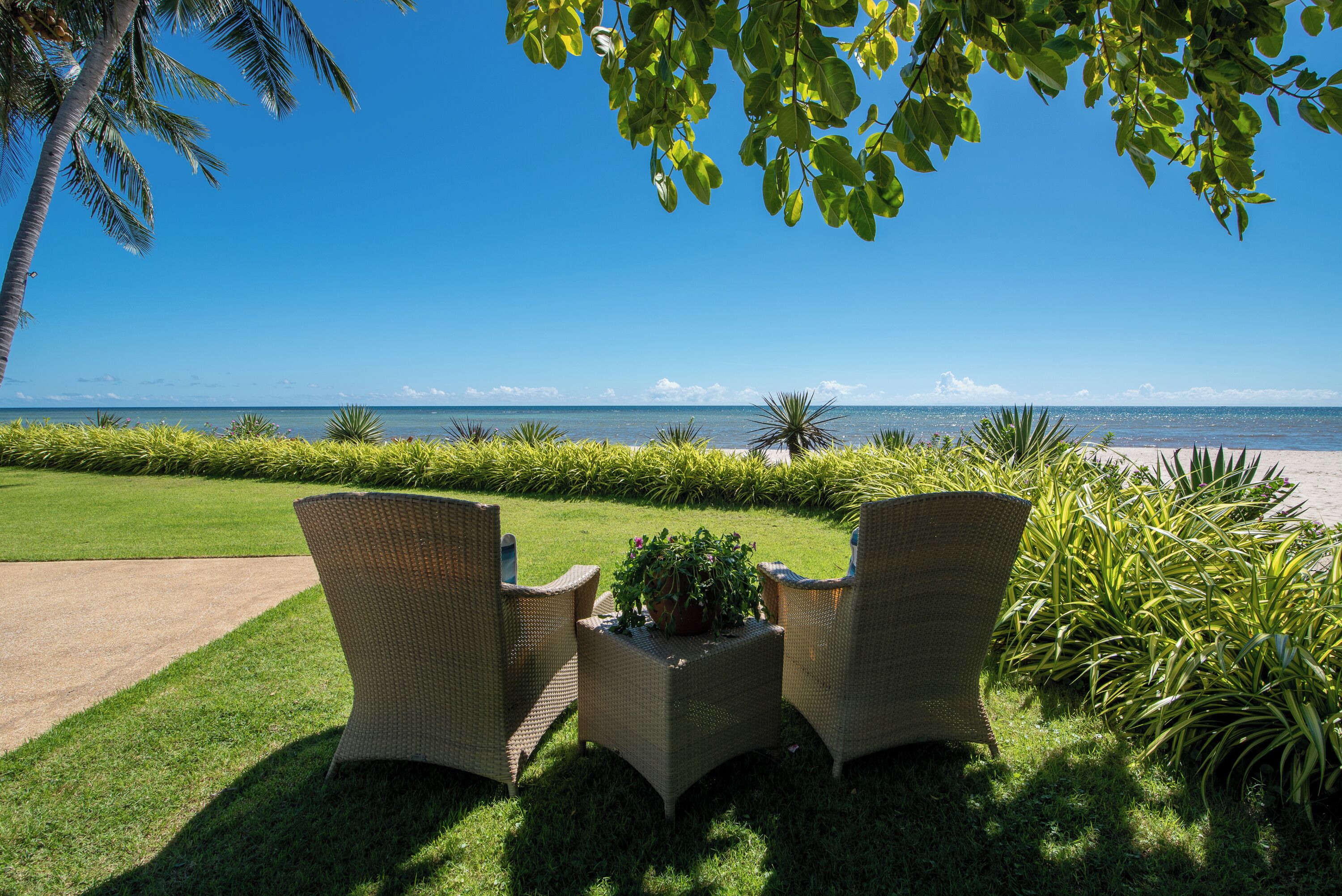 Une terrasse avec des fauteuils en osier et une petite table agrémentée d’une plante, d’où l’on a vue sur une plage et l’océan.