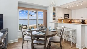 A dining area with a view of the sea, a kitchen with white cabinets, and a dining table set with plates and glasses.