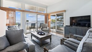 A spacious living area with a sofa, armchairs, a coffee table, and a TV. There is a balcony with a view of the water.