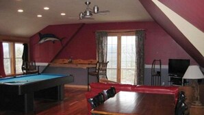 Ein Zimmer mit Poolbillard, einem Holzespeisetisch mit Stühlen, einem Deckenventilator und einem großen Fenster mit Vorhängen.