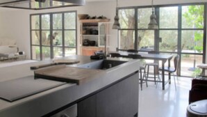 Una cocina moderna con una isla grande, un comedor con sillas y vistas a la vegetación a través de amplios ventanales.