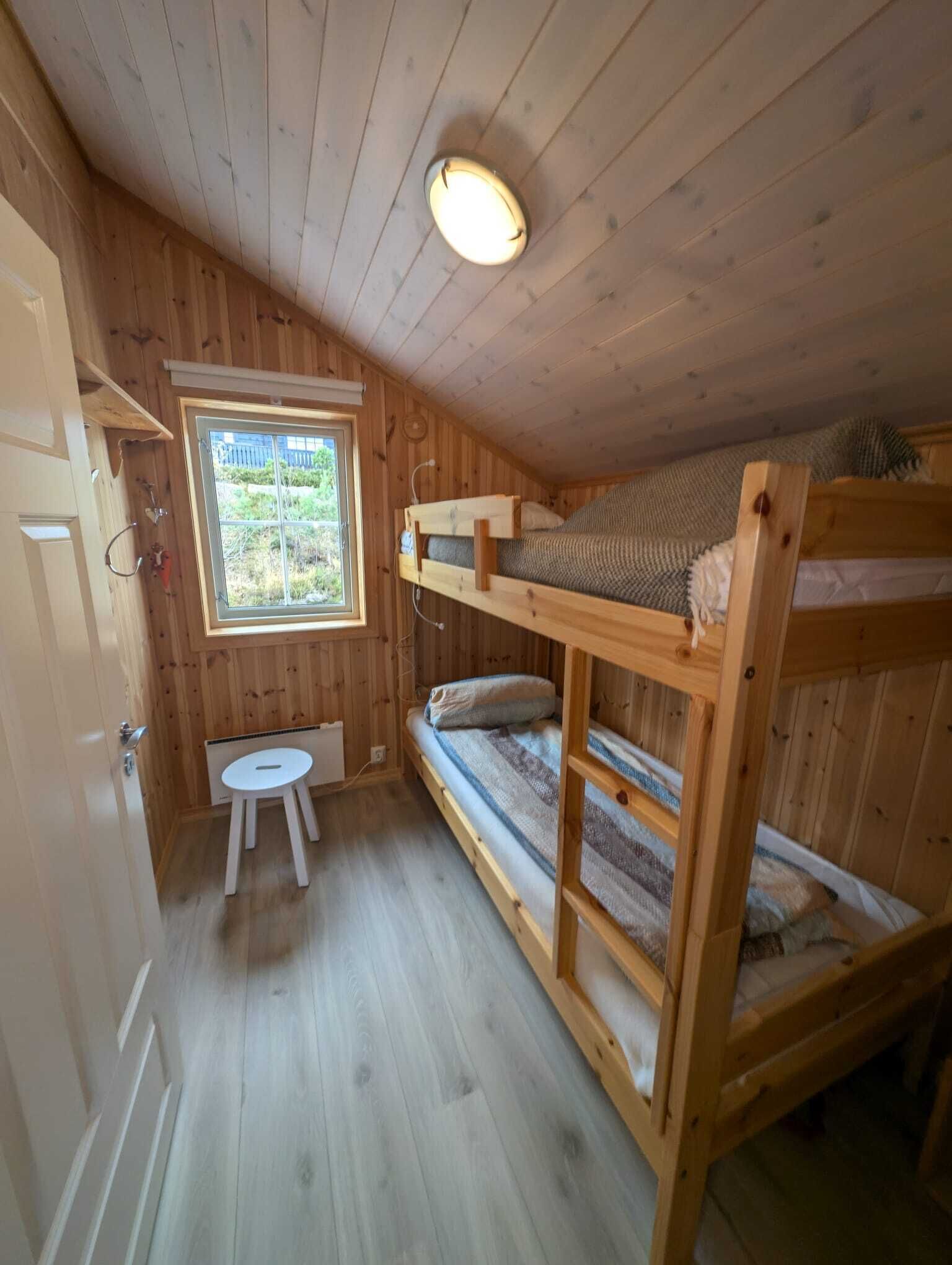 A small, wooden cabin room with bunk beds, a small table, and a window with a view of greenery.