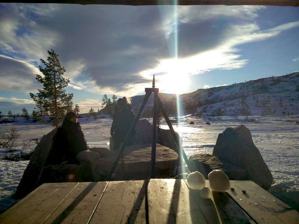 Puisesta pöydästä näkyy pari valkoista kenkää, metallinen jalusta ja lumipeitteinen maisema, jonka yllä on kirkas taivas.
