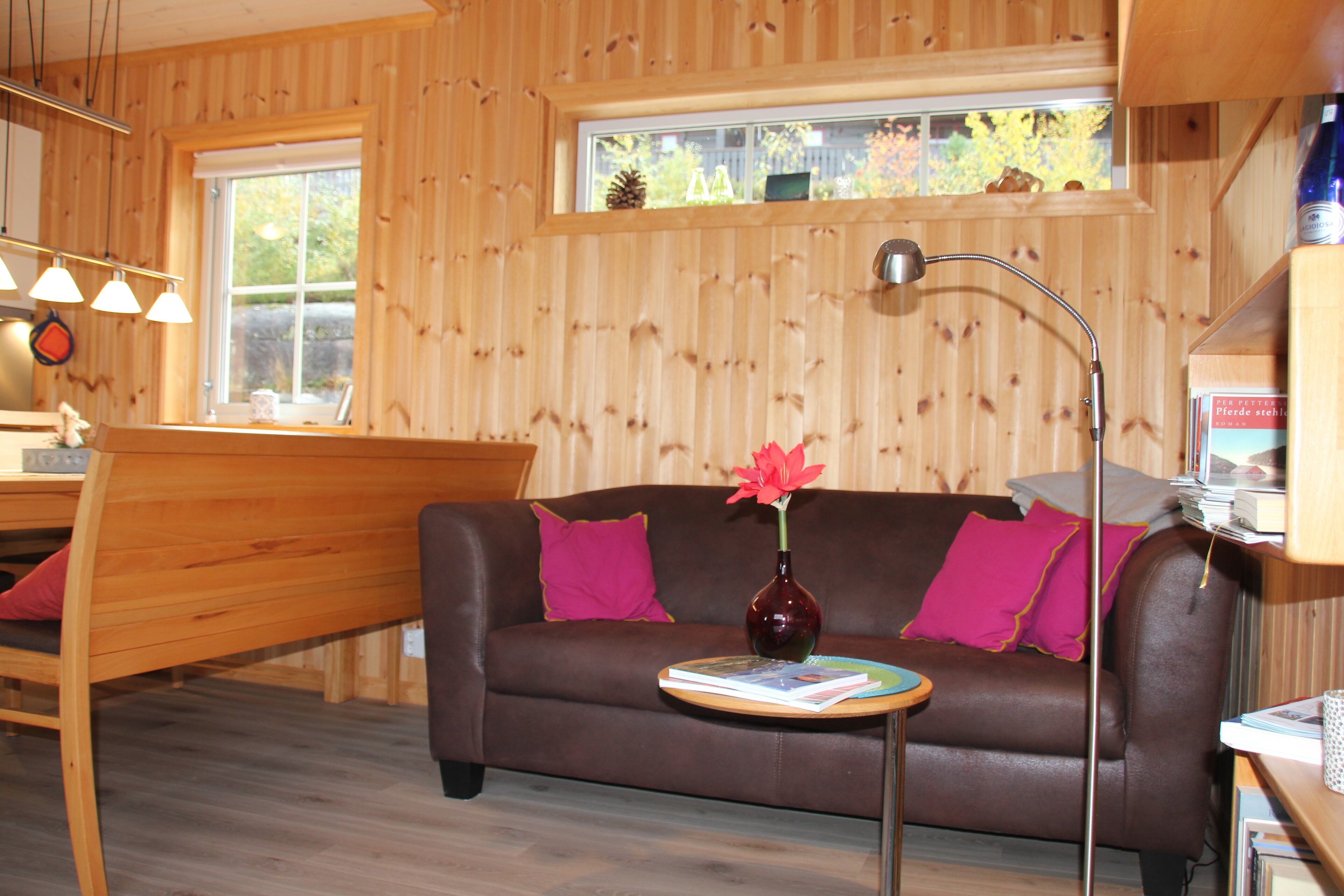 Ein gemütliches Zimmer mit einem braunen Ledersofa, einem kleinen Holztisch mit einer Vase und Büchern sowie einem Fenster mit Blick auf Bäume.
