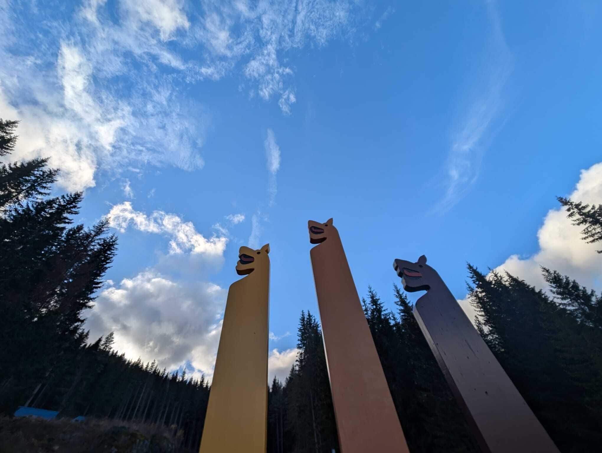 Tres esculturas altas y estilizadas con rostros de animales, frente a un cielo azul despejado y un bosque frondoso.