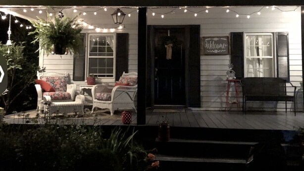 A porch with string lights, two white wicker chairs, a red polka dot cushion, and a 'Welcome' sign.