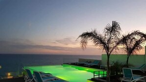 Een dakterras zwembad met oneindige rand, groene verlichting, lounge stoelen en uitzicht op de oceaan bij zonsondergang.
