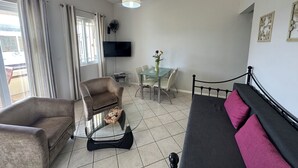 Un salon moderne comprenant un canapé, un fauteuil, une table basse en verre, une table à manger et des chaises. Un téléviseur à écran plat est fixé au mur, et un ventilateur de plafond est visible.