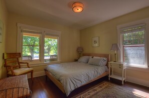 A bedroom with a bed, two bedside tables, a chair, a small table, and two windows.