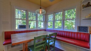Un comedor con mesa de madera y bancos acolchados rojos, ventanas amplias con vistas a la vegetación y una lámpara colgante.
