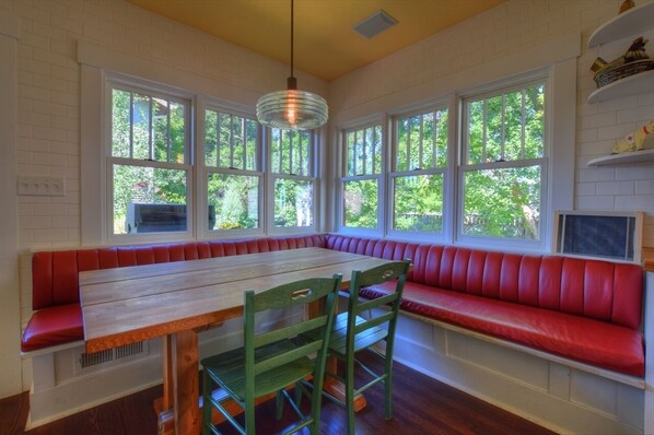 A dining area with a wooden table and red cushioned bench seating, large windows with a view of greenery, and a hanging light fixture.