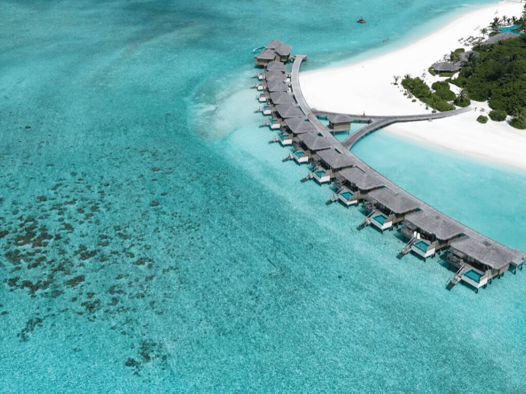 Vista aérea de un complejo turístico con bungalows sobre el agua, conectados por una pasarela curva.