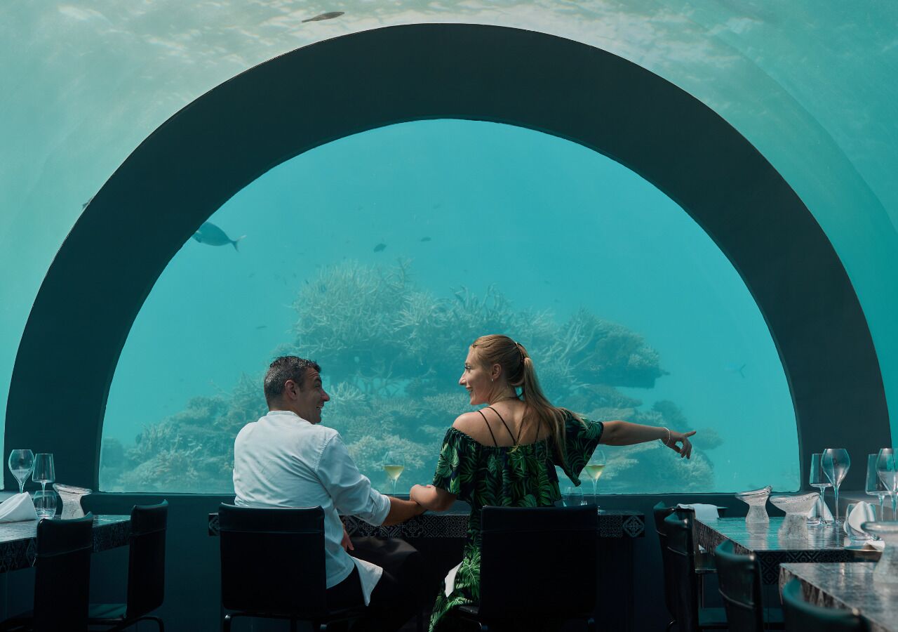 Un couple est assis à une table près d’une grande fenêtre en arc d’où l’on peut admirer un paysage sous-marin.