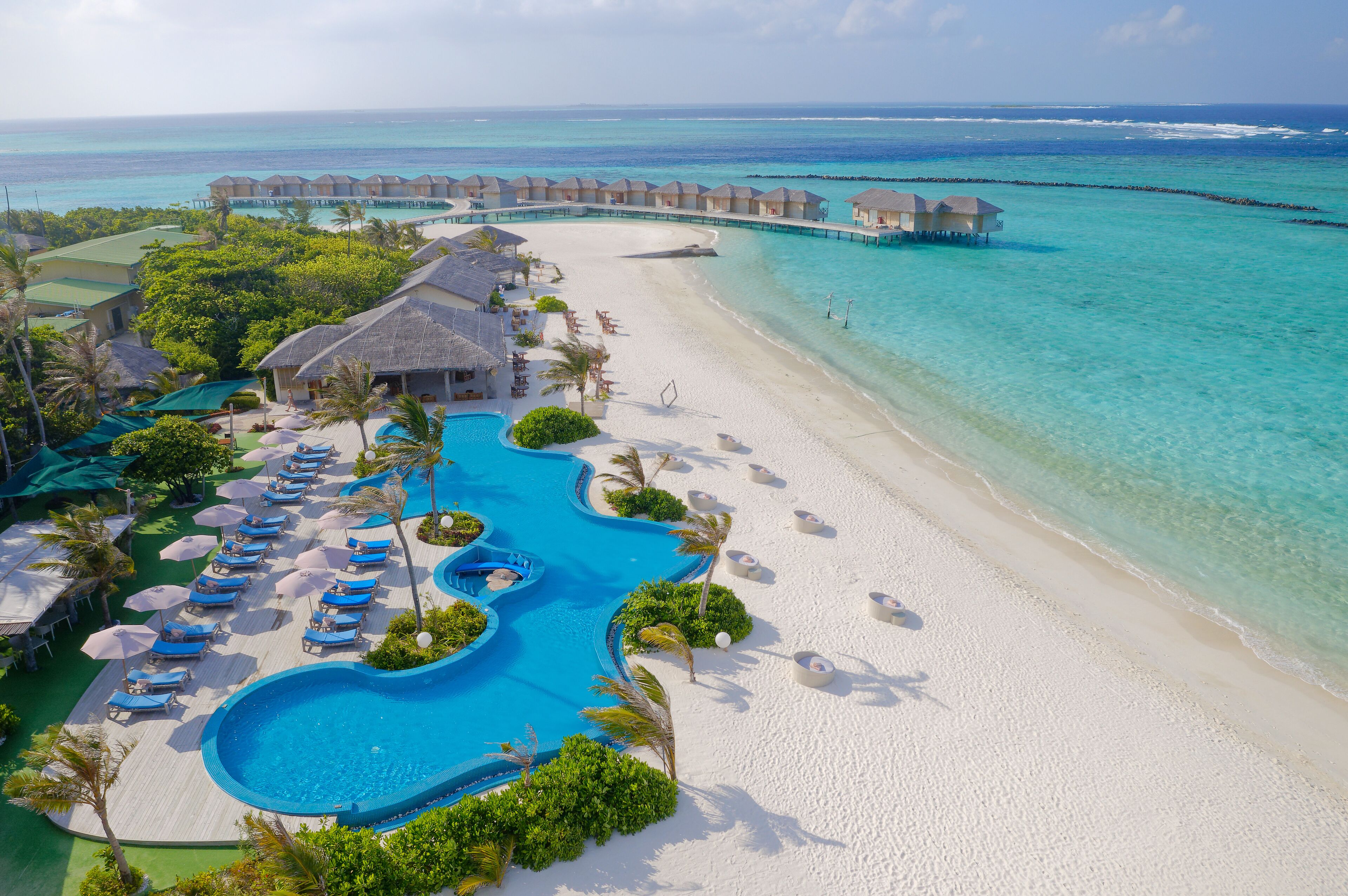 Luchtbeeld van een resort met een groot zwembad, strand en bungalows op palen.
