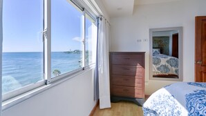 Ett sovrum med ett stort fönster med utsikt över havet, en byrå i trä och en säng med mönstrat sänglinne.