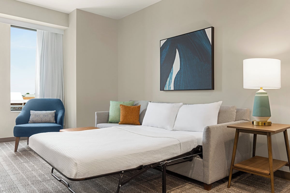 A hotel room with a bed, a chair, a sofa, a lamp, and a painting on the wall.