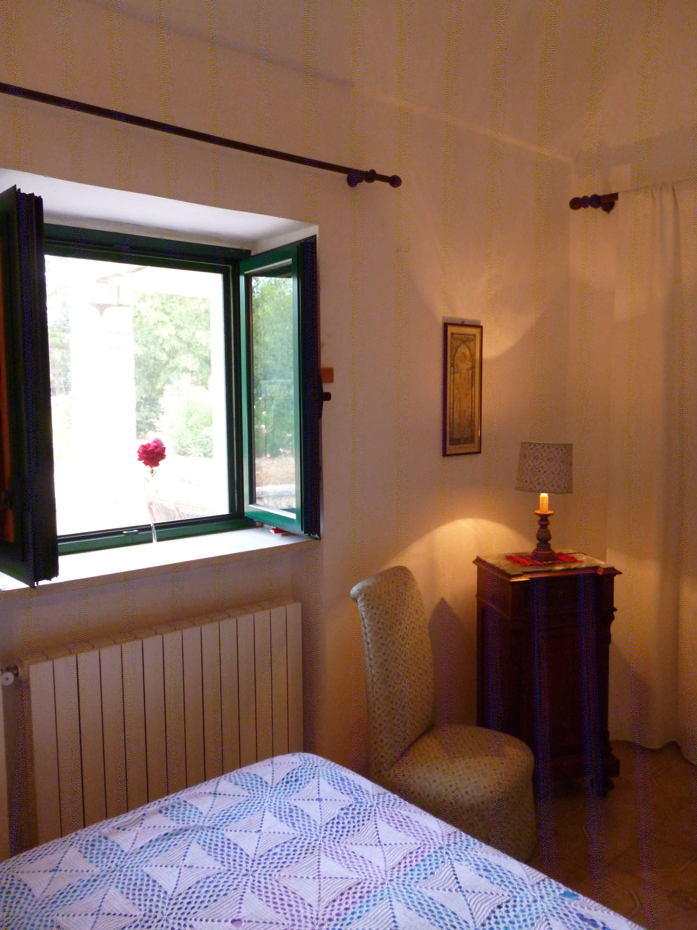 Une chambre à coucher avec un lit, une chaise, une table avec une bougie, une fenêtre avec des rideaux et un radiateur.