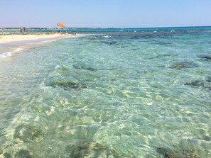Een strand met helder ondiep water, een zandstrand en een uitzicht op een stad in de verte.