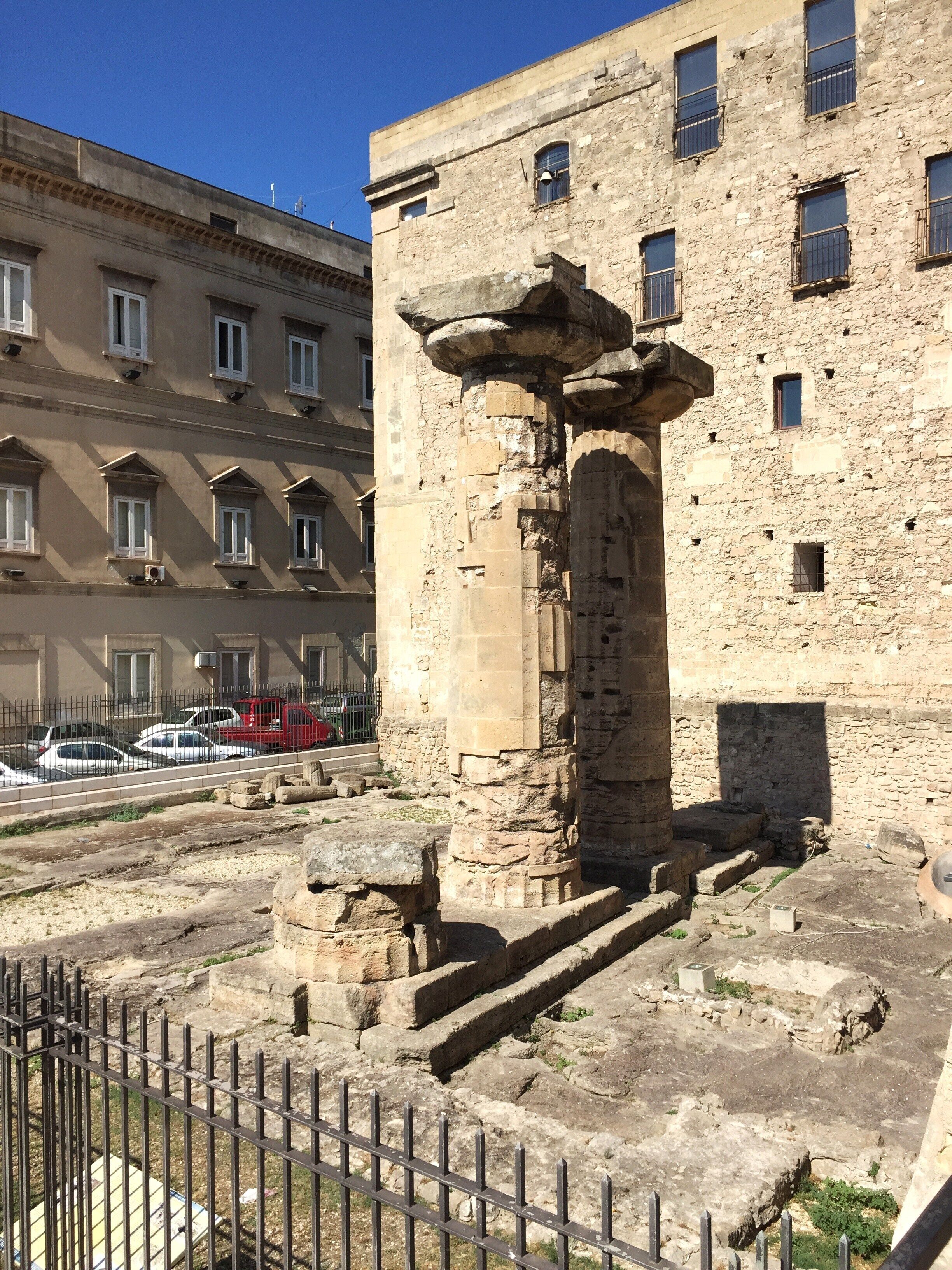 Ein historisches Steingebäude mit Säulen und Stufen, umgeben von einem Metallzaun, in einer städtischen Umgebung mit modernen Gebäuden im Hintergrund.