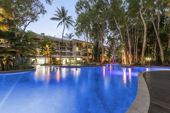 A large, illuminated swimming pool surrounded by trees and a hotel building.