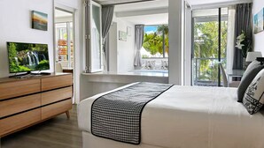 A bedroom with a large bed, a TV on a wooden dresser, and a view of a balcony with plants and a window. - Lxry 2 bed Apt-Sea and Mountain View (Palm Cove)