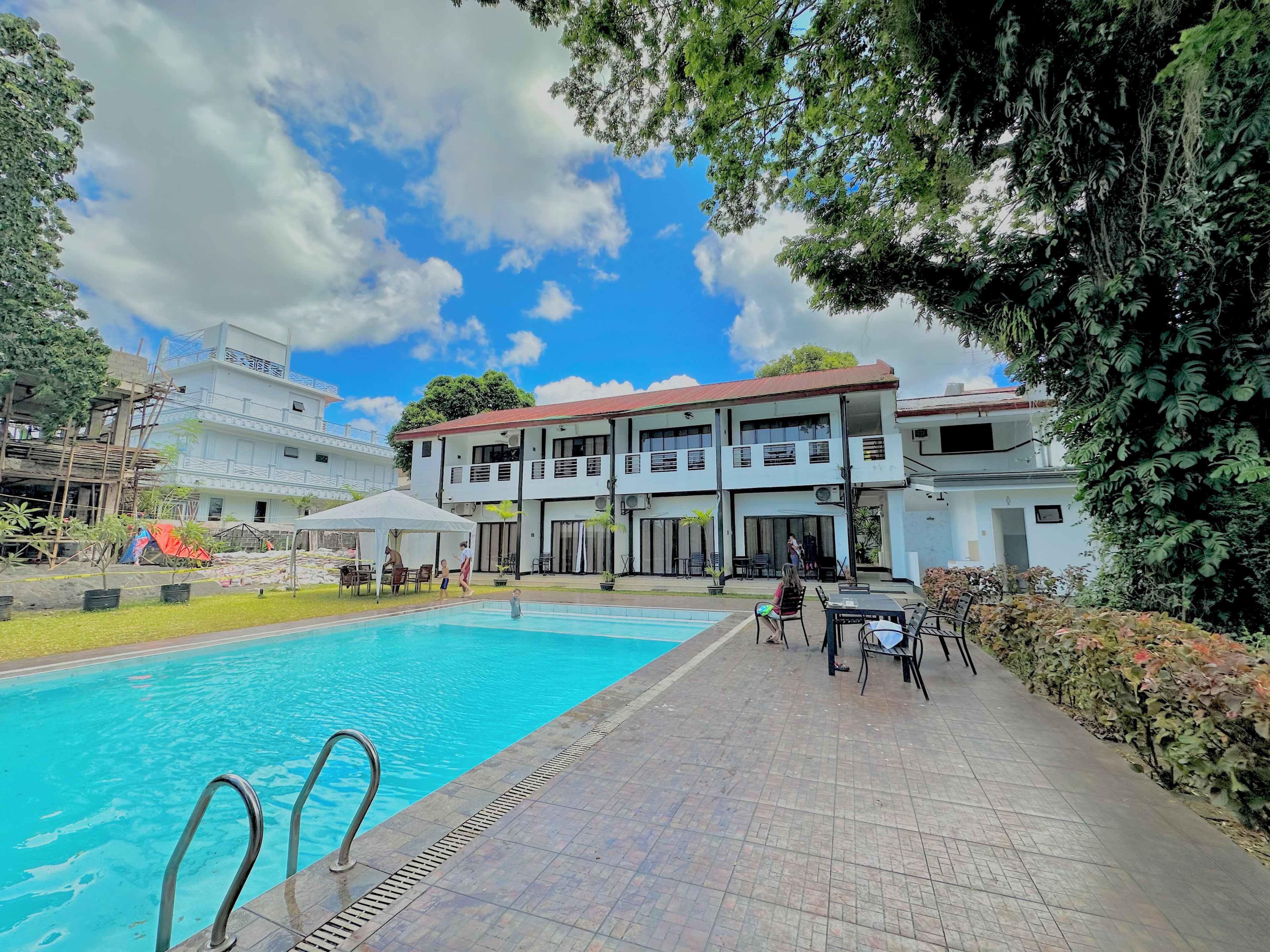 A swimming pool with a tiled deck, a covered seating area, and a two-story building with balconies.