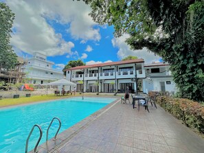 Una piscina con plataforma de azulejos, una zona de estar cubierta y un edificio de dos pisos con balcones.