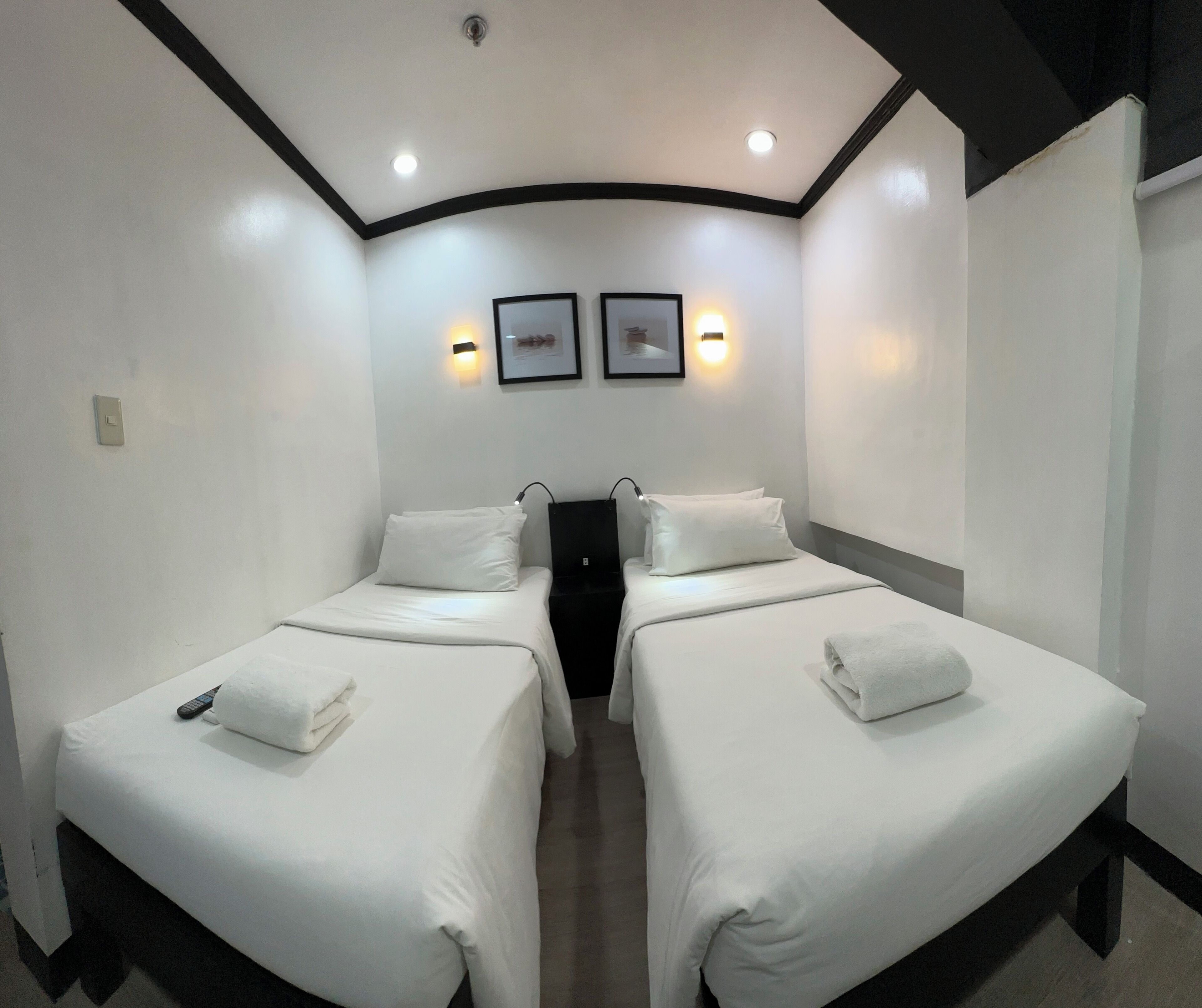 A hotel room with two single beds, each with a white bedspread and pillows. There are wall-mounted light fixtures above the beds and a light switch on the wall.