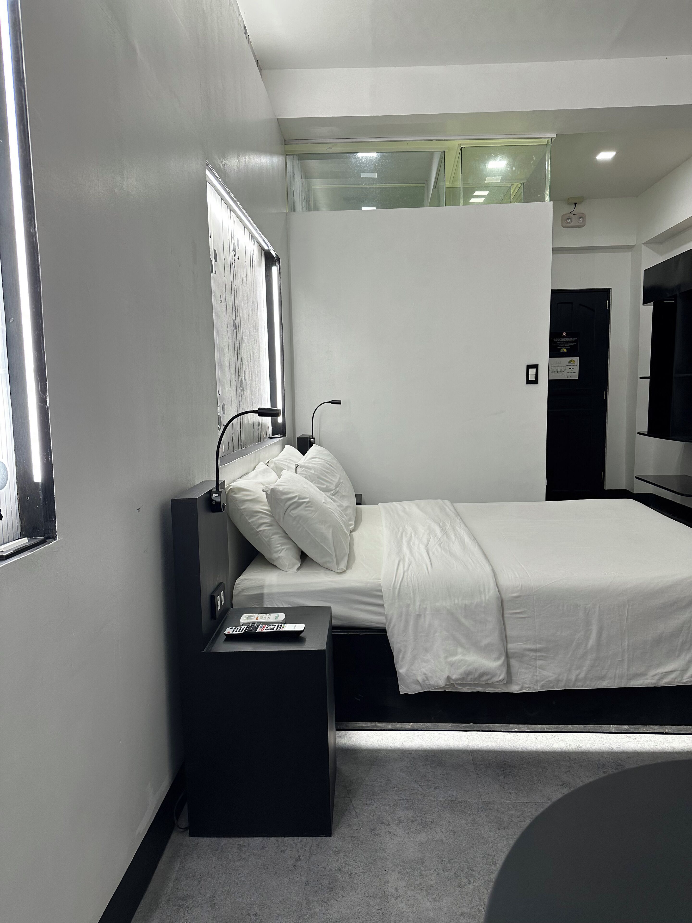 Una habitación de hotel moderna con una cama individual, una mesita de noche pequeña, una lámpara de noche, un televisor y una ventana con persianas.