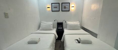 Ein Hotelzimmer mit zwei Einzelbetten, jeweils mit Kissen und einem gefalteten Handtuch. Es gibt zwei Wandleuchten und einen kleinen Tisch mit einer Fernbedienung.