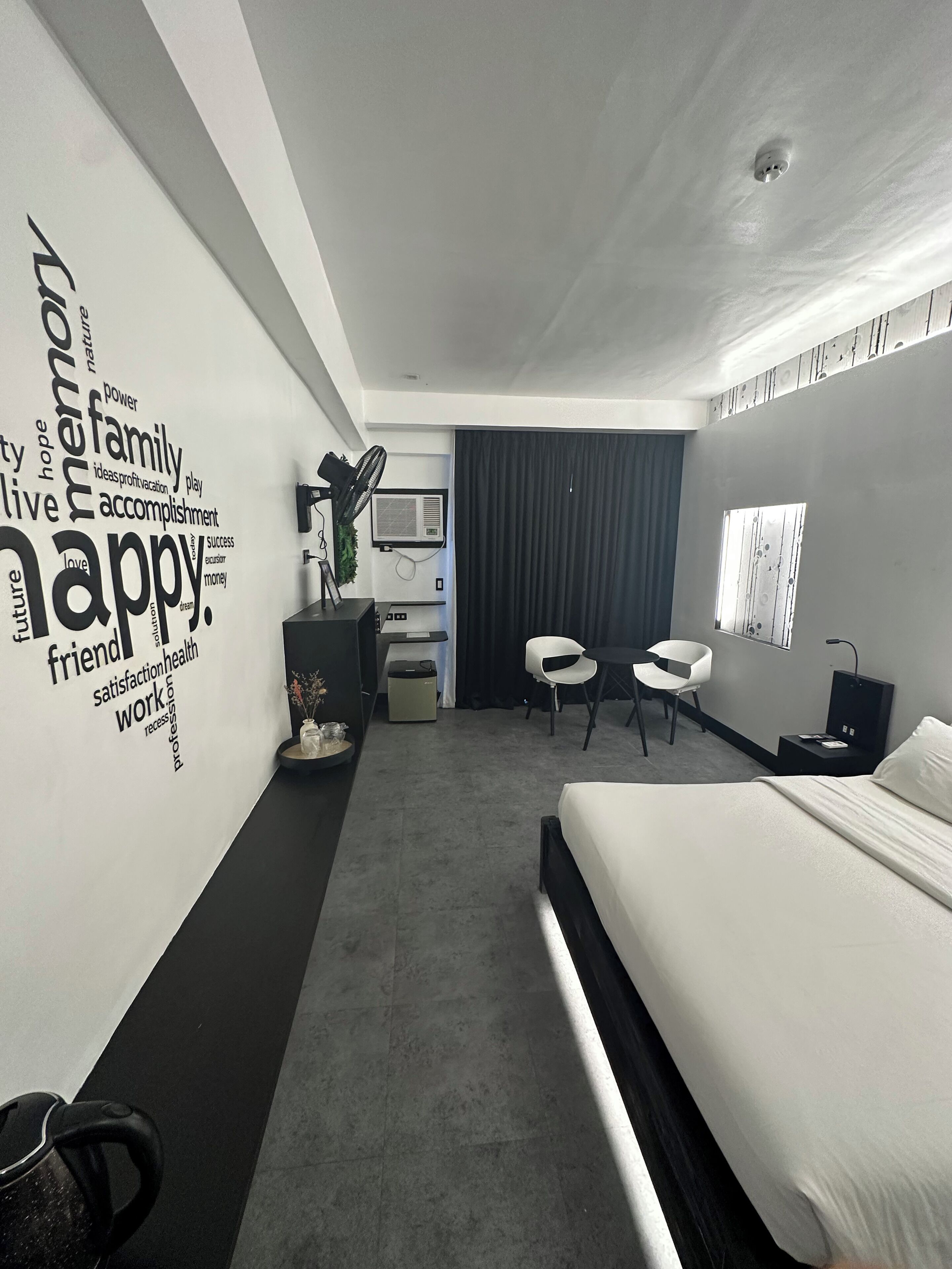 A modern hotel room with a large bed, a desk, two chairs, a small table, and a wall with motivational words.