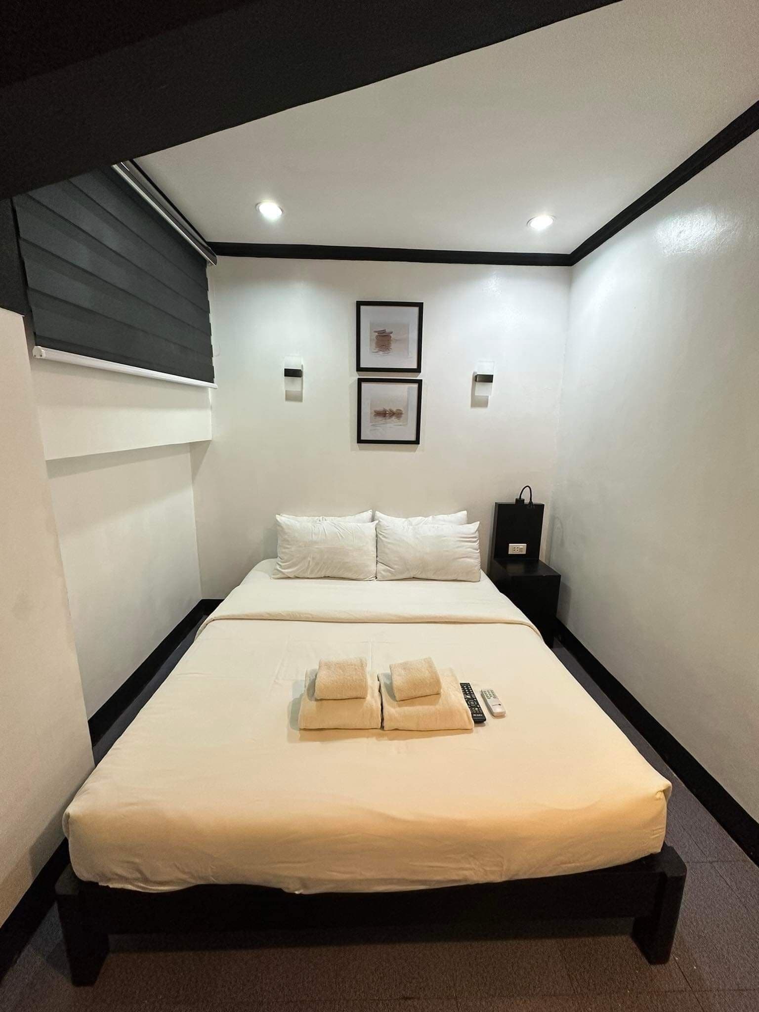 A hotel room with a bed, pillows, a bedside table, a remote control, and two framed pictures on the wall.