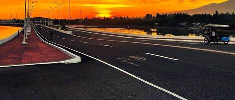 Un pont avec une passerelle piétonne rouge et des lampadaires, qui mène à un point de vue sur un coucher de soleil sur l’eau.