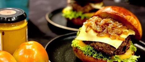 A burger with cheese and caramelized onions on a bun, accompanied by a jar of mustard, tomatoes, and garlic.