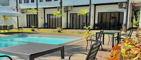 Zona de piscina con sillones y mesas, rodeada por un edificio de varios pisos y balcones.