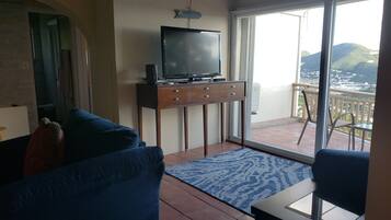 A living room with a blue sofa, a TV on a wooden stand, and a balcony with a view of the city.