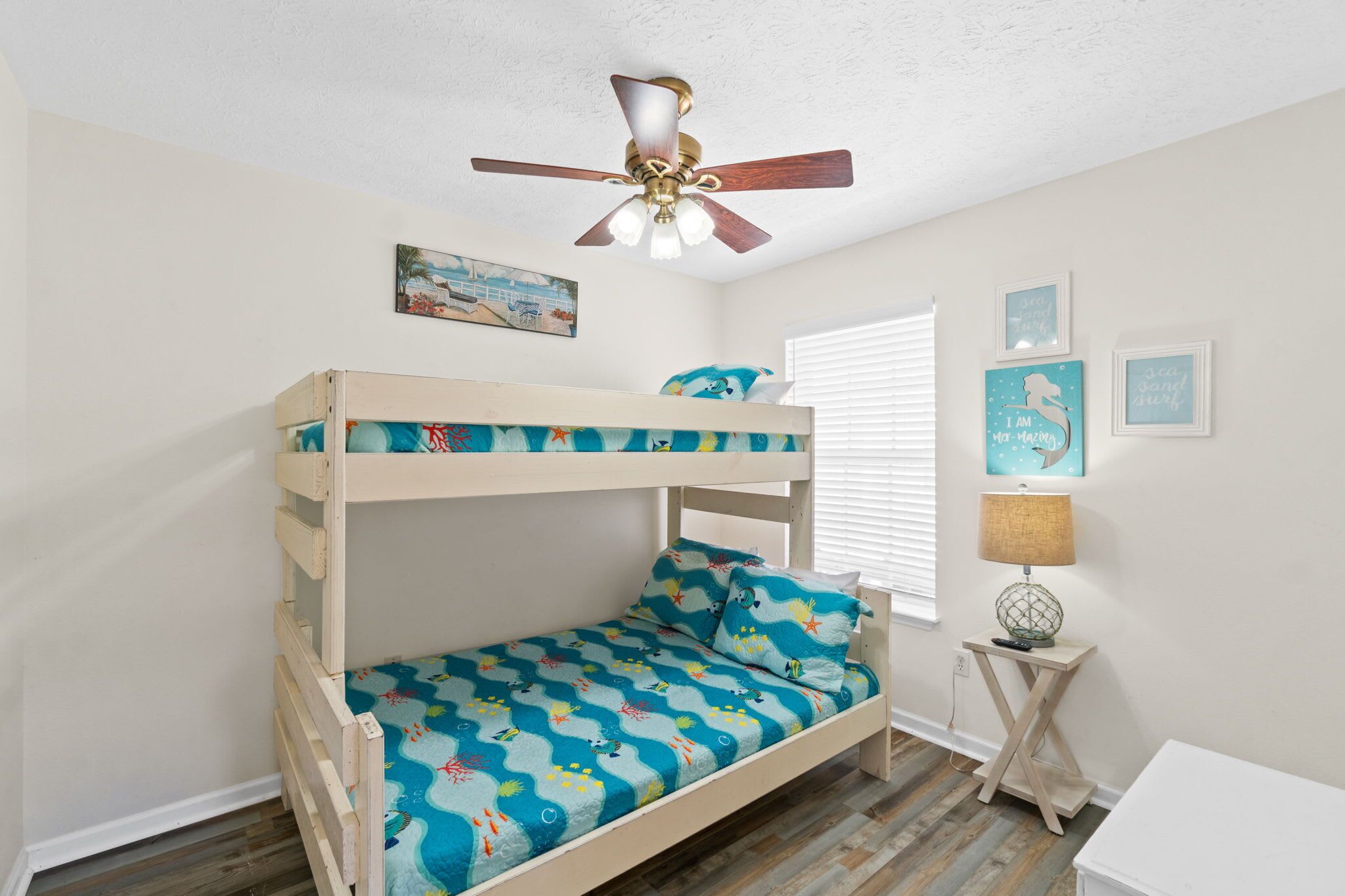 A bunk bed with a blue ocean-themed bedspread, a ceiling fan, and a small table with a lamp.