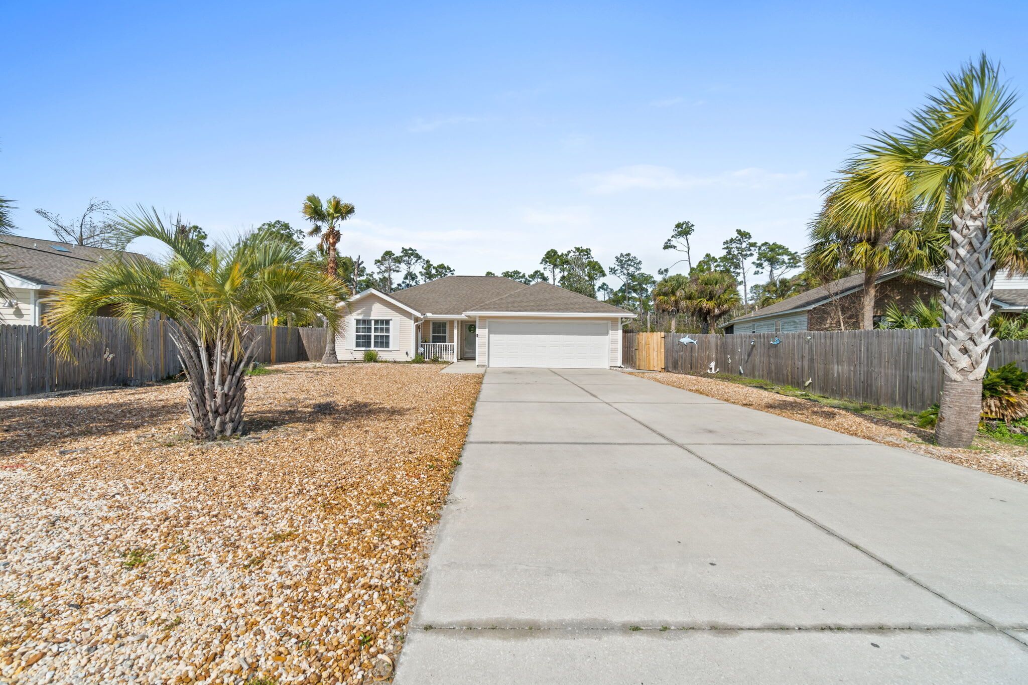 A single-story house with a garage, surrounded by a wooden fence and palm trees.