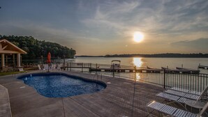 A pool area with a view of the sunset over a lake, surrounded by lounge chairs and a wooden building.