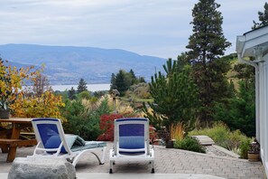 A patio with two lounge chairs, a wooden table, and a view of mountains and a lake. - Dreams B & B - Rock Creek Room (Summerland)