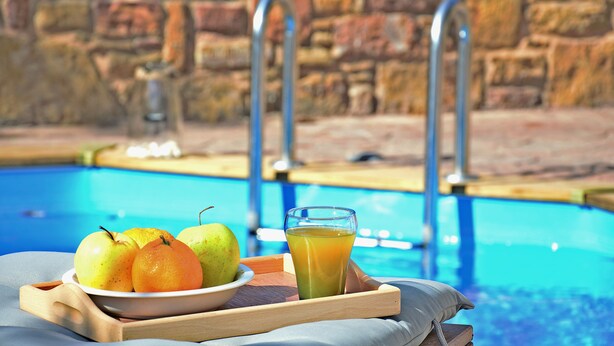 A tray with fruits and a glass of juice by a poolside with stone walls. - Mastiha Villas in Town (Chios)