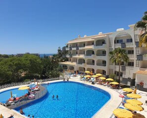 A hotel with a large swimming pool surrounded by sun loungers and umbrellas, with a view of the sea.