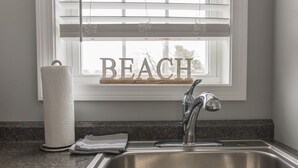 Eine Küche mit Spüle aus Edelstahl, einem Papierhandtuchhalter und einem Fenster mit dem Schriftzug 'BEACH'.