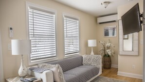 Ein Wohnzimmer mit grauem Sofa, zwei Fenstern mit Jalousien, einem an der Wand montierten Fernseher, einer Lampe und einer Vase mit Blumen.