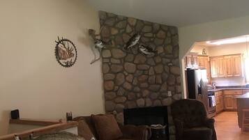 A living room with a stone fireplace, a ceiling fan, and a kitchen in the background.