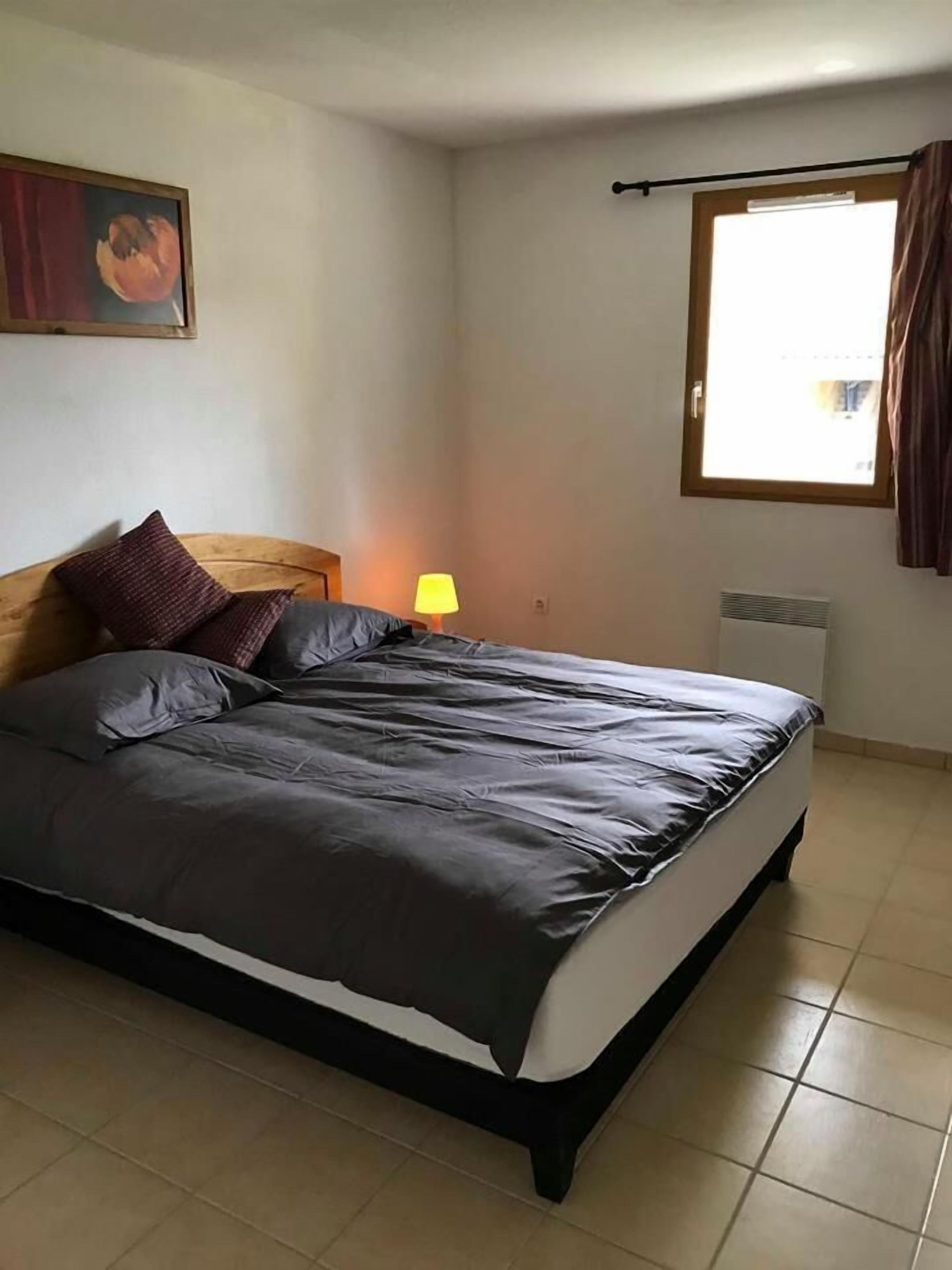 Quarto com cama, mesa de cabeceira, abajur, radiador, janela e um quadro na parede.