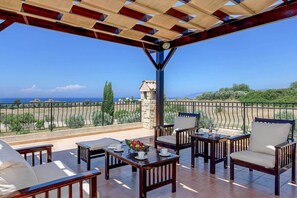 A covered outdoor seating area with a view of the sea, furnished with wooden chairs and tables, and a stone structure in the background. - Villa Yialos (Neo Chorio)