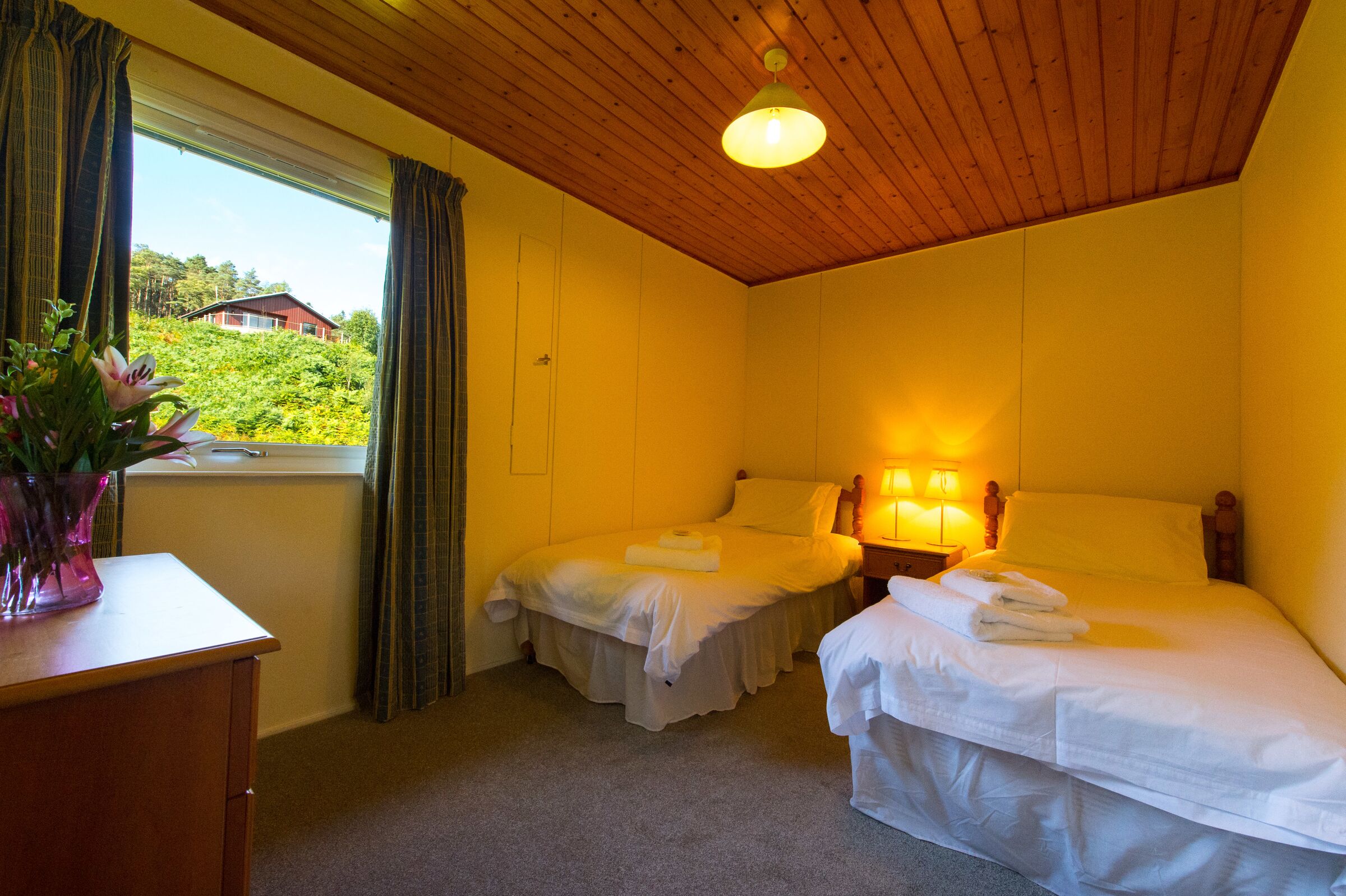 A hotel room with two beds, a wooden ceiling, a window with curtains, and a bedside lamp.