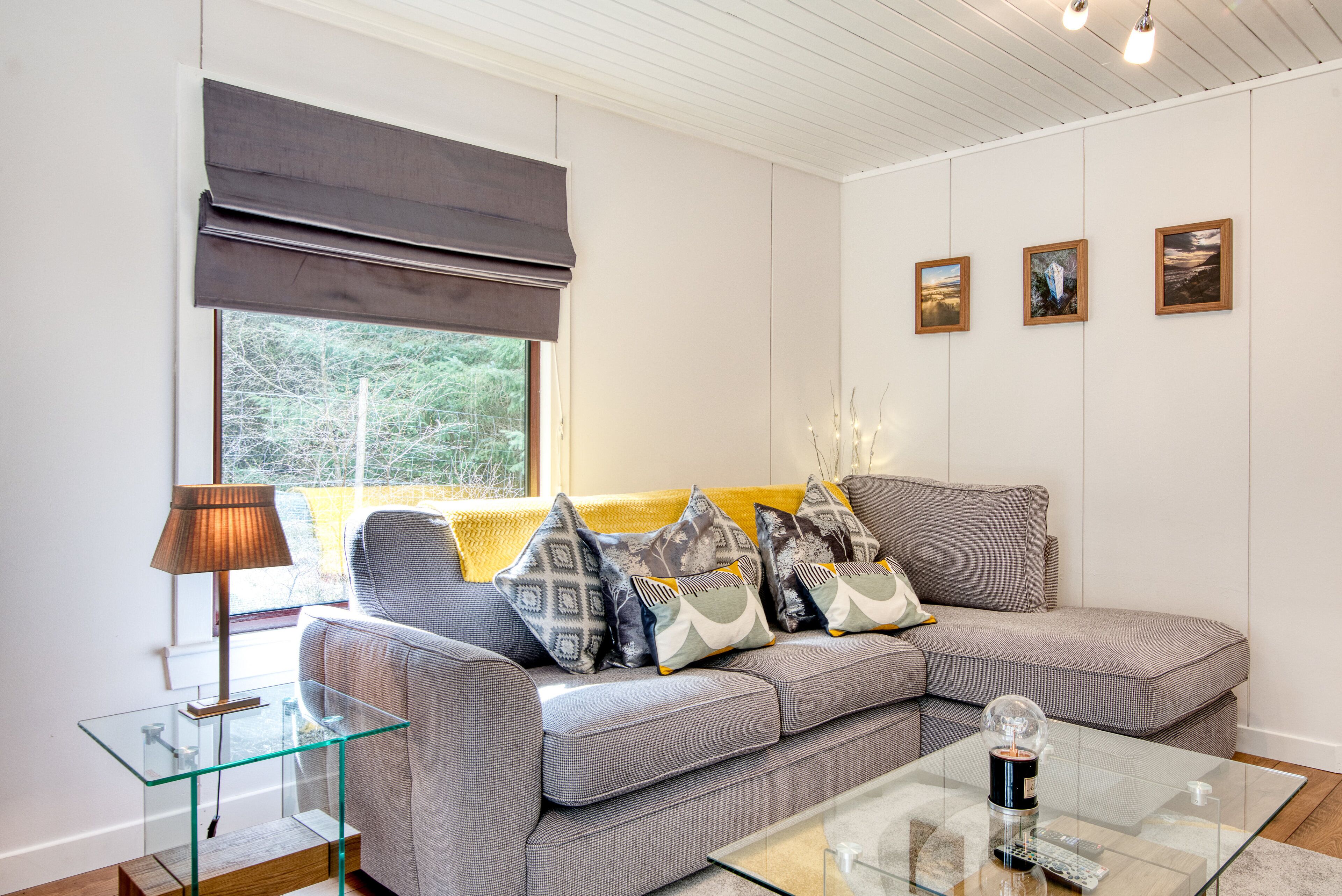 A modern living room with a grey sectional sofa, a glass coffee table, and a large window with a grey roller blind. There are three framed pictures on the wall and a lamp on a glass side table.
