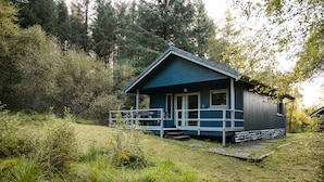 A small, single-story cabin with a porch and a front door, surrounded by dense forest. - Otter With Mountain View and Enclosed Decking (Crianlarich)